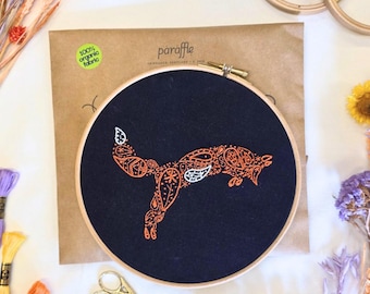 Könnte beinhalten: Ein Stickrahmen mit marineblauem Stoff und einem orangefarbenen Fuchsmotiv. Der Fuchs ist mit einem Paisley-Muster verziert. Das Set enthält eine braune Papierverpackung mit dem Text "paraffle" und "100% organic fabric".