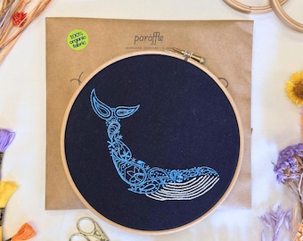 以下が含まれることがあります： 濃紺の生地に、水色のクジラのデザインが施された刺繍枠。クジラはペイズリーと花の模様で装飾されています。木製の枠は、ドライフラワーや刺繍用品（ハサミなど）に囲まれています。