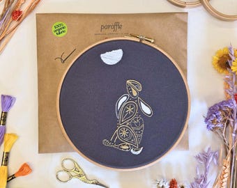 May include: An embroidery kit featuring a navy blue fabric with a gold and white rabbit design. The kit includes a wooden hoop, embroidery floss, and a brown paper package with the text "100% organic fabric" and the brand name "paraffle".