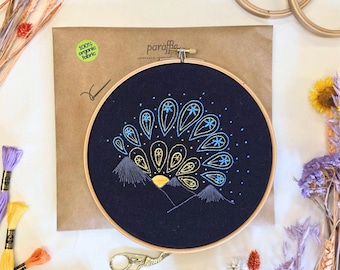 May include: An embroidery hoop with a navy blue fabric featuring a colorful design of a sunrise over mountains. The design is stitched with yellow, blue, and gray thread. The hoop is displayed with embroidery floss, scissors, and dried flowers.