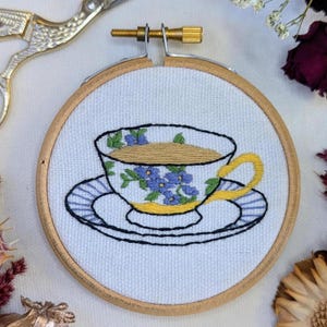 May include: A hand-stitched embroidery hoop displaying a teacup and saucer design. The teacup is decorated with blue flowers and a yellow handle, set against a white background. The saucer has blue stripes. The embroidery is framed in a wooden hoop.