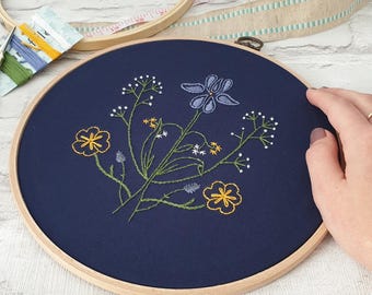 Può includere: Un cerchio da ricamo blu navy con un motivo floreale. Il disegno presenta fiori gialli e blu con steli e foglie verdi.