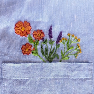 May include: A light blue linen fabric with embroidered flowers in shades of orange, purple, and green. The flowers are arranged in a cluster and are centered on the fabric.