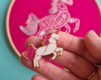 May include: A gold-tone enamel pin featuring a white unicorn with a pink mane and tail. The unicorn is standing on all four legs with its head held high.