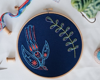 Könnte beinhalten: Ein runder Stickrahmen mit einem marineblauen Stoff, der ein Vogel- und Blattmuster zeigt. Der Vogel ist mit blauem und rotem Faden umrandet, mit orangefarbenen Sternakzenten. Das Blattmuster ist grün und gelb. Holzspulen und bunte Fäden sind sichtbar.