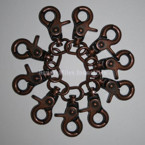 Puede incluir: Conjunto de mosquetones giratorios de color bronce dispuestos en círculo. Cada gancho está conectado a un pequeño eslabón de cadena, creando un diseño decorativo. El texto "SquareMiles Industries" es visible.