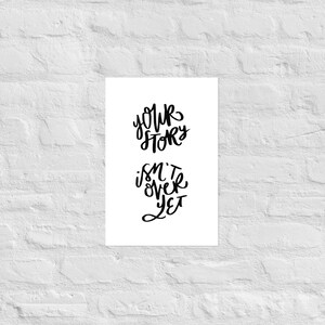 May include: A white poster with black handwritten text that reads "Your Story Isn't Over Yet." The text is arranged in two lines, with "Your Story" above "Isn't Over Yet." The poster is mounted on a white brick wall.