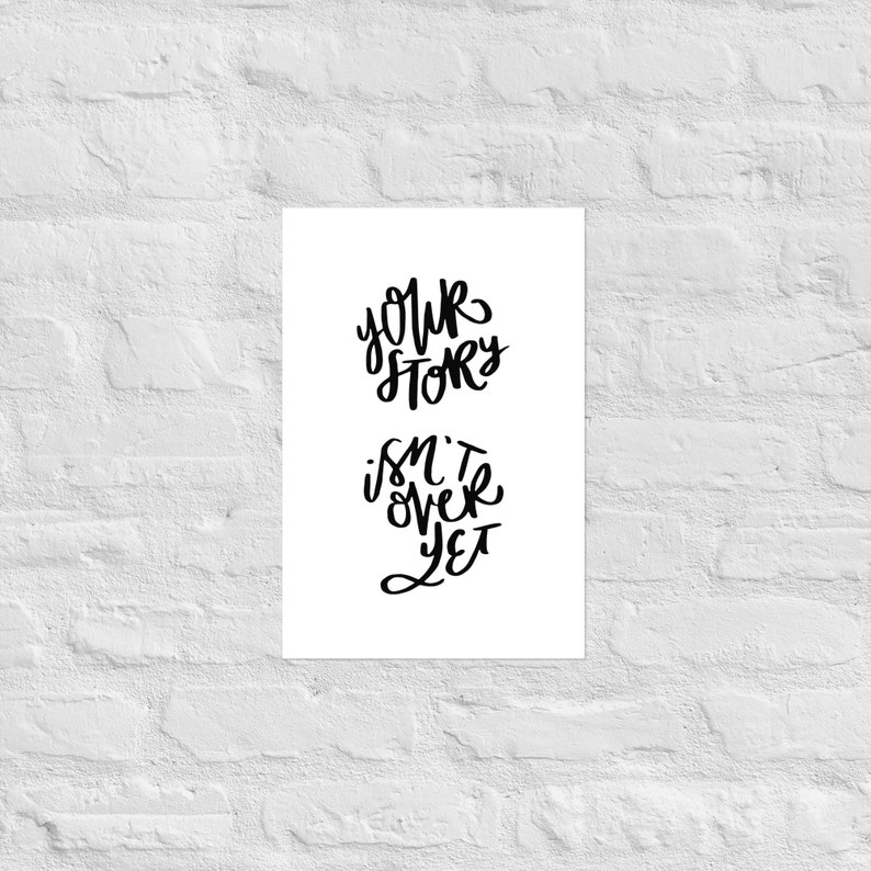 May include: A white poster with black handwritten text that reads "Your Story Isn't Over Yet." The text is arranged in two lines, with "Your Story" above "Isn't Over Yet." The poster is mounted on a white brick wall.