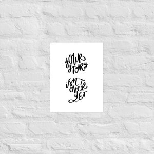 May include: A white print with black lettering against a white brick wall. The text reads "YOUR STORY ISN'T OVER YET" in a handwritten style. The print is rectangular and centered.
