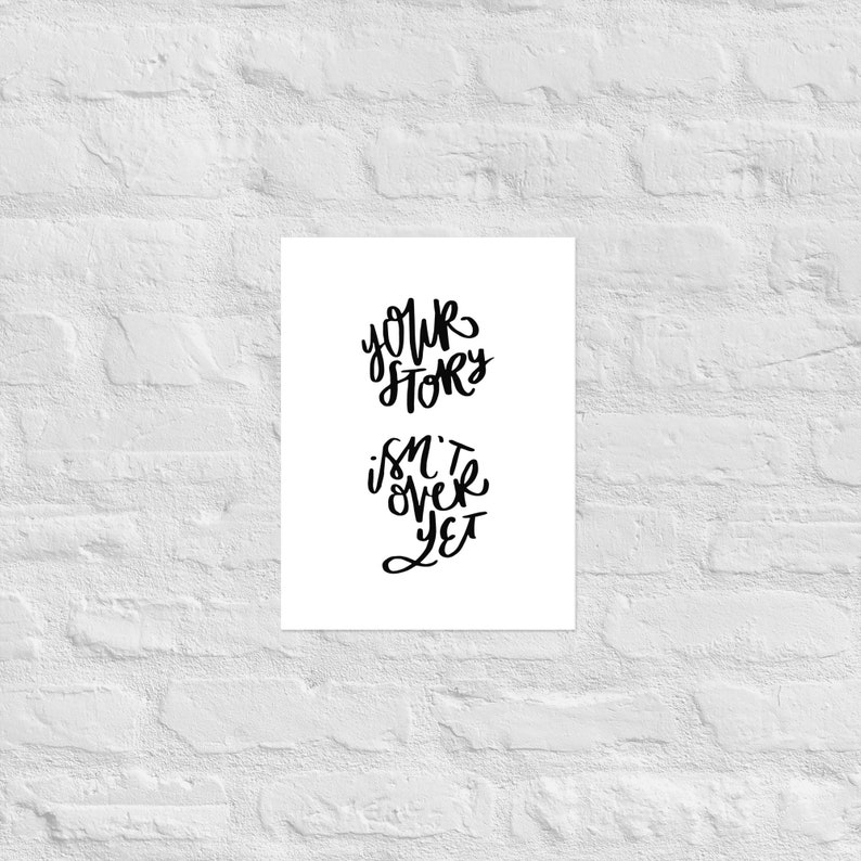 May include: A white print with black lettering against a white brick wall. The text reads "YOUR STORY ISN'T OVER YET" in a handwritten style. The print is rectangular and centered.