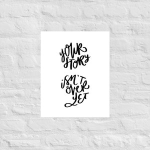 May include: A white poster with black cursive text that reads "Your Story Isn't Over Yet." The text is arranged in two lines, with "Your Story" above "Isn't Over Yet." The poster is displayed against a white brick wall.