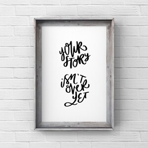 May include: A framed print with the words "Your Story Isn't Over Yet" in black script. The print is set in a gray wooden frame and hangs against a white brick wall.