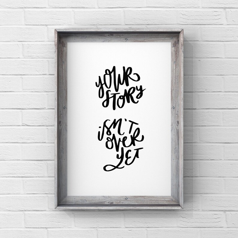 May include: A framed print with the words "Your Story Isn't Over Yet" in black script. The print is set in a gray wooden frame and hangs against a white brick wall.