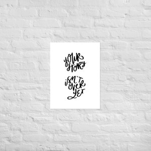 May include: A white print with black lettering that reads "Your story isn't over yet." The text is in a handwritten style. The print is displayed against a white brick wall.