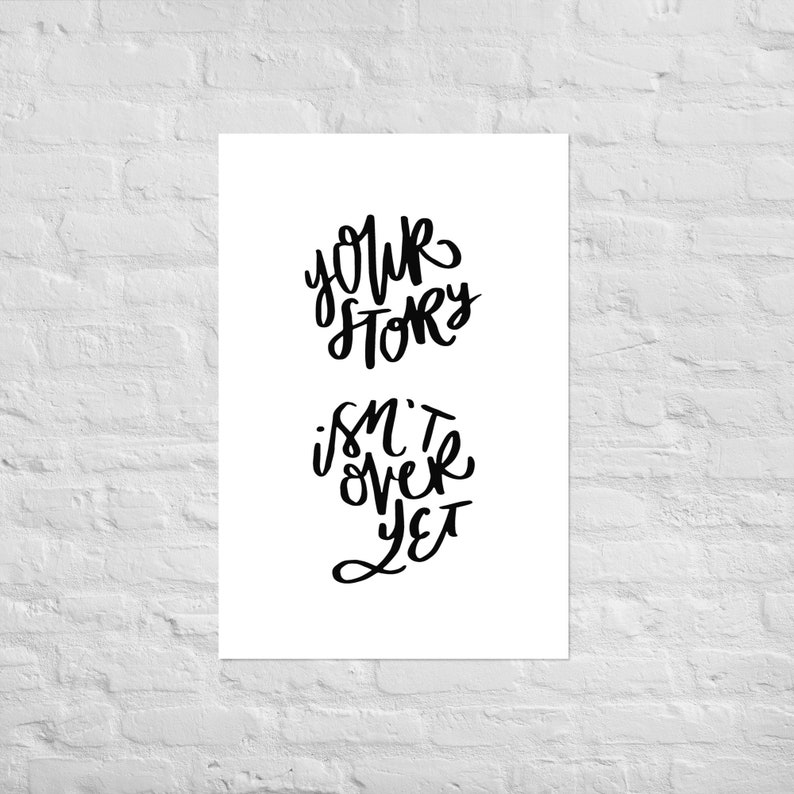 May include: White poster with black handwritten text that reads "Your Story Isn't Over Yet". The words are in a cursive font. The poster is displayed against a white brick wall.