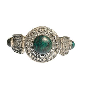 May include: Silver-toned bracelet featuring a large, round, dark green stone at the center. The bracelet has intricate, detailed patterns and two smaller green stones on the sides. The design is ornate and eye-catching.
