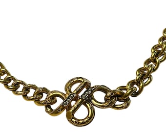 May include: A gold-toned necklace with a detailed chain and a central decorative clasp. The clasp features an intricate design with small, embedded gemstones. The necklace is set against a white background, highlighting its ornate details.