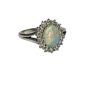 Può includere: Anello d'argento con un'opale ovale circondata da piccole gemme trasparenti. L'opale mostra colori iridescenti. Il gambo dell'anello si divide in due alle spalle.