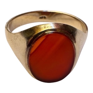 May include: A gold signet ring featuring a smooth, oval-shaped, deep orange carnelian stone set into the center. The band is polished gold with a rounded profile, showcasing a classic design with a solid metal shank and a bezel-set gemstone.
