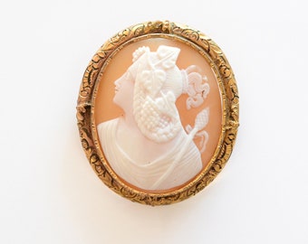 May include: A gold-toned cameo brooch featuring a carved portrait of a woman with a floral headdress. The cameo is set in a detailed oval frame.