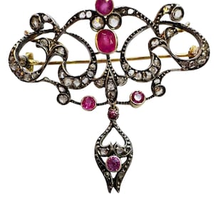 May include: An ornate brooch featuring a dark metal filigree frame set with multiple small clear stones and several vibrant pink oval and round gemstones. The design includes a central vertical drop element and a horizontal pin mechanism on the back.
