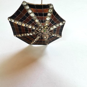 May include: A vintage brooch in the shape of a small umbrella. The umbrella has a black and brown checkered pattern with a spider and web design. The spider has a green gem for its eye and is surrounded by clear rhinestones.
