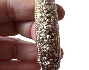 May include: A gold-toned metal bangle bracelet featuring an intricate embossed floral pattern with raised petals and swirling vine details. The edges of the bracelet are lined with a fine beaded texture, creating a decorative border along the entire length.