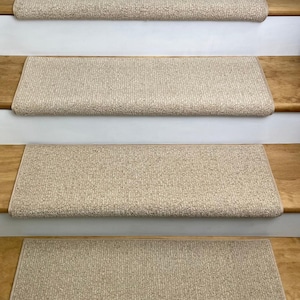 May include: Four beige stair treads with a textured, woven appearance are installed on wooden stairs. The treads are rectangular with rounded edges, providing a non-slip surface. The stairs have white risers and wooden steps.
