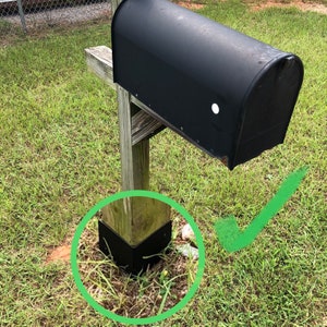 May include: A black mailbox mounted on a wooden post with a black plastic base. The post is surrounded by green grass.
