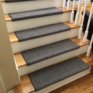 May include: A set of gray stair treads installed on wooden stairs. The treads are rectangular with a dark gray border and a textured surface. The stairs have white risers and a wooden handrail.