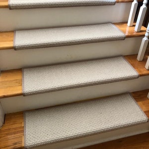 May include: Four stair treads with a light beige, textured surface and a darker beige border are installed on wooden stairs. The stairs have white risers and a wooden handrail.