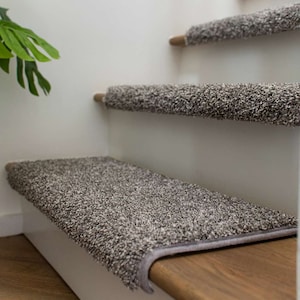 May include: Close-up of a set of gray carpet stair treads installed on wooden stairs. The treads have a textured surface and are designed to provide traction and prevent slipping.