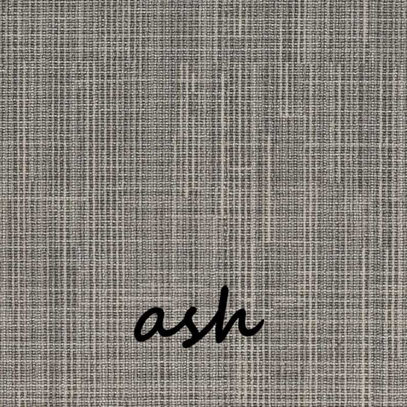 May include: Close-up of a textured fabric in shades of gray and off-white. The word "ash" is written in a cursive font across the bottom right corner of the fabric. The fabric has a woven appearance.