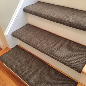 May include: A set of brown stair treads installed on wooden stairs. The treads have a woven texture and a dark brown border. The stairs are painted white and the flooring is hardwood.