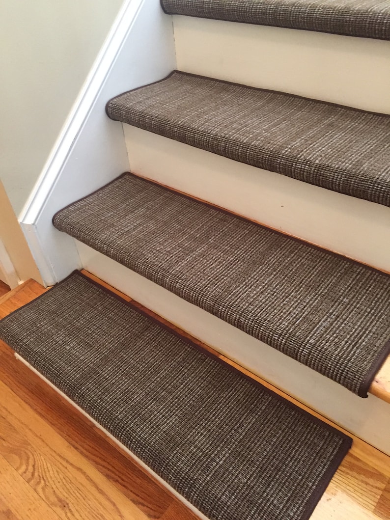 May include: A set of brown stair treads installed on wooden stairs. The treads have a woven texture and a dark brown border. The stairs are painted white and the flooring is hardwood.