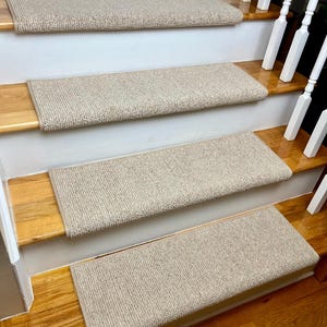 May include: Beige carpet stair treads with a textured surface. The treads are installed on wooden stairs with a natural wood finish.