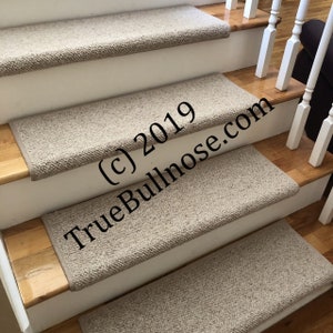 May include: Beige carpet stair treads with a textured surface. The treads are installed on a set of wooden stairs with white painted risers. The text "(c) 2019 TrueBullnose.com" is visible on the treads.