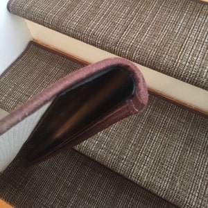 May include: Brown and beige stair treads with a woven pattern. The edge of the tread is a darker brown, creating a border. The treads are installed on wooden stairs.