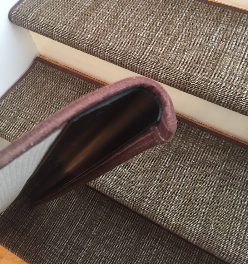 May include: Brown and beige stair treads with a woven pattern. The edge of the tread is a darker brown, creating a border. The treads are installed on wooden stairs.