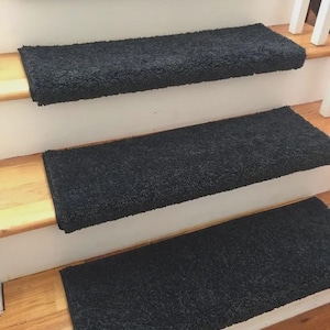 May include: Four black, plush stair treads are installed on wooden stairs. The treads are rectangular and cover the stair steps. The stairs have white risers and a wooden handrail is visible in the background. The treads add a decorative and safety element.