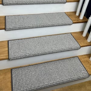 May include: Stair treads with a gray and white woven pattern are installed on wooden stairs. The treads have a dark gray border and are rectangular. The stairs have white risers and a wooden handrail.