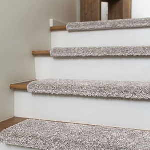 May include: Close-up of a set of beige carpet stair treads on a white staircase. The treads are made of a soft, plush material and have a subtle texture. The treads are designed to add a touch of warmth and comfort to any staircase.