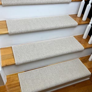 May include: Beige carpet stair treads with a textured woven pattern. The treads are installed on wooden stairs with white risers.