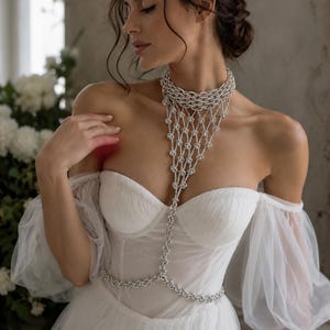 Puede incluir: Un vestido blanco de hombros descubiertos con mangas abullonadas transparentes y escote coraz&oacute;n, combinado con un collar corporal plateado que presenta una gargantilla de varias capas y un dise&ntilde;o drapeado tipo red hasta la cintura.