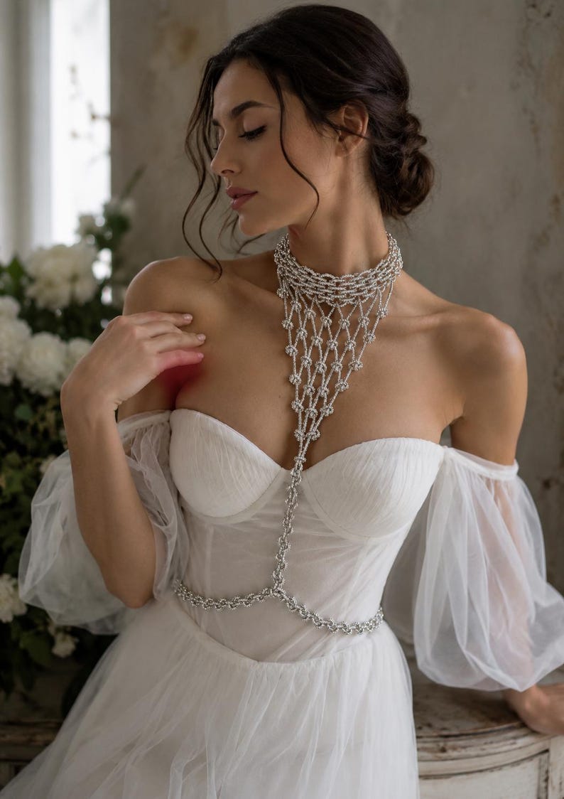 Puede incluir: Un vestido blanco de hombros descubiertos con mangas abullonadas transparentes y escote coraz&oacute;n, combinado con un collar corporal plateado que presenta una gargantilla de varias capas y un dise&ntilde;o drapeado tipo red hasta la cintura.