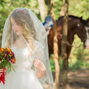 May include: A bride wearing a white wedding dress and veil, holding a bouquet of red and orange flowers. She is standing in a forest setting with a horse in the background.
