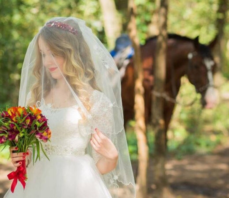 May include: A bride wearing a white wedding dress and veil, holding a bouquet of red and orange flowers. She is standing in a forest setting with a horse in the background.