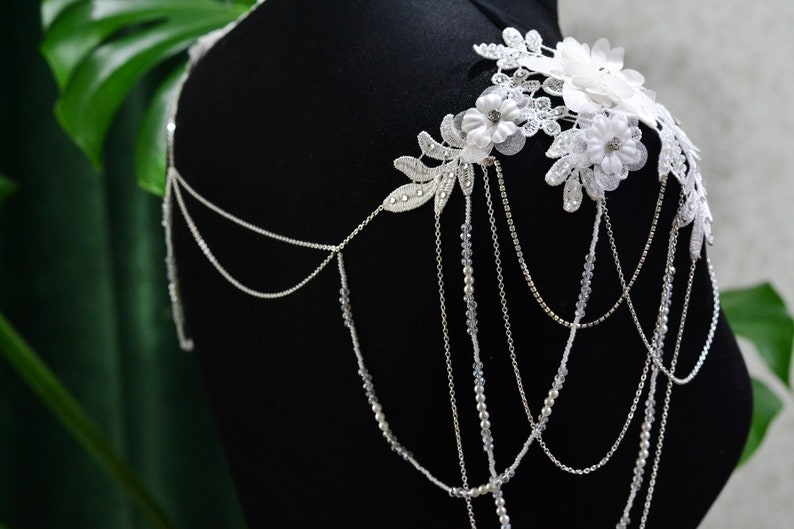 May include: A white lace and pearl bridal shoulder piece with a floral design. The piece is made of delicate lace and features a variety of pearls and beads.