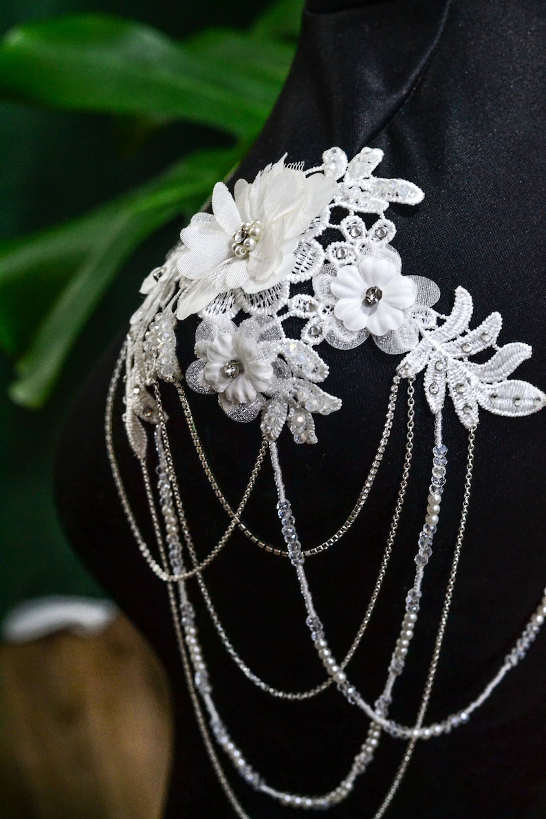 May include: White lace shoulder piece with delicate floral embellishments and a silver chain with crystal beads.
