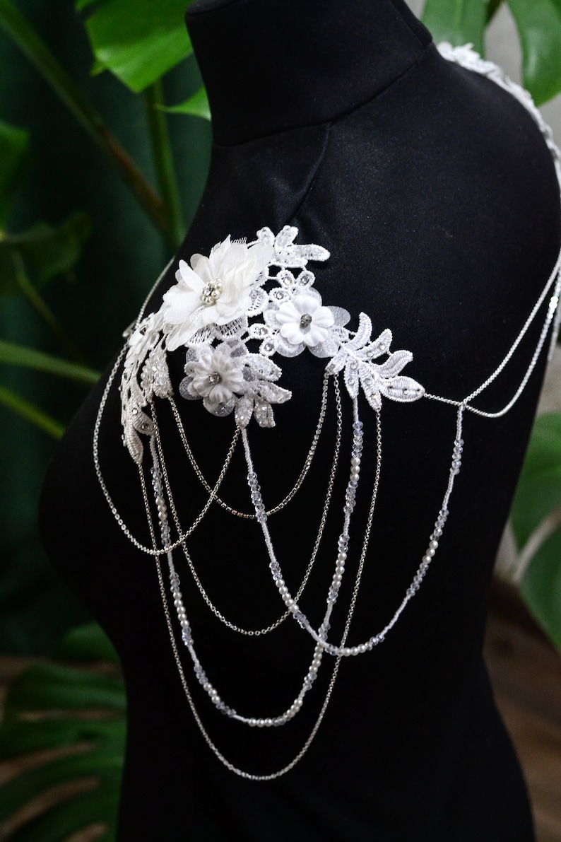 May include: A white lace shoulder piece with delicate floral accents and a cascading chain of silver and pearl beads.